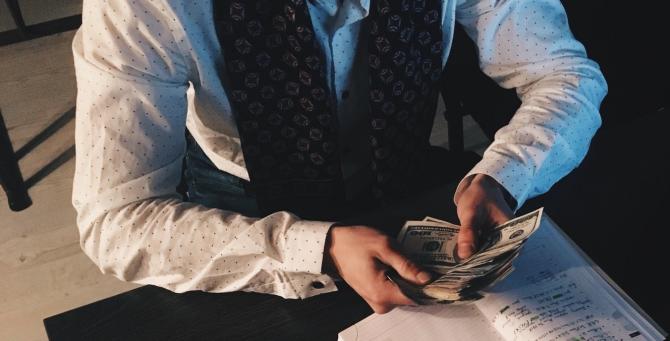 person in dress shirt and vest counting a stack of money at a table, in front of an open notebook