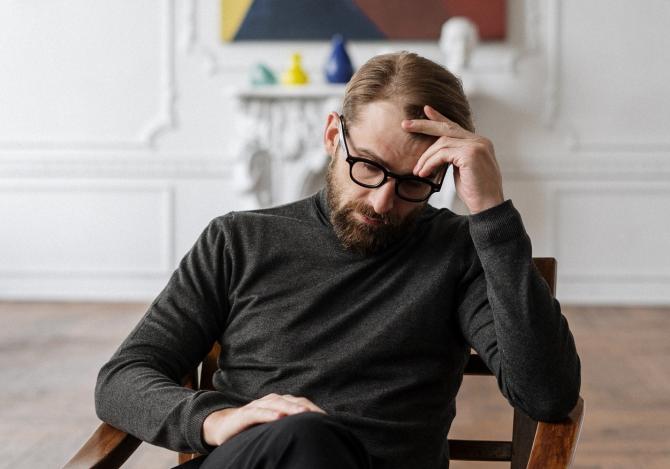 bearded man with glasses, clothed in black, sitting in chair leaning on one elbow, hand to forehead, with worried or sad expression