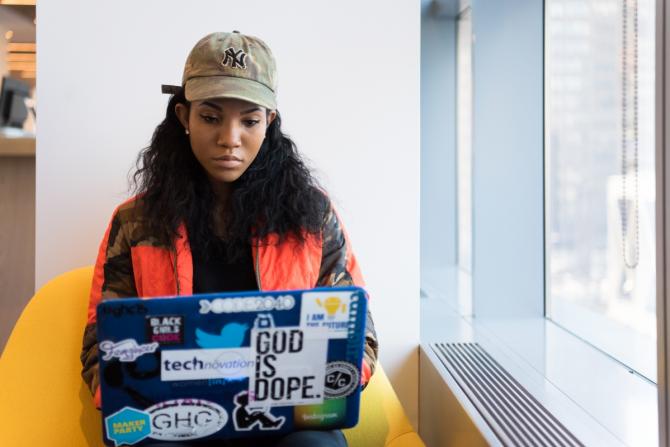 Black woman seated in yellow chair typing in laptop festooned with stickers - some read "God is dope," "Black Girls Code", "Femgineer", "I am the future"