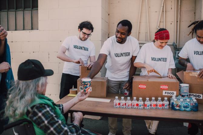 multiracial group of volunteers handing out food and water to man in wheelchair with long gray hair, flannel shirt, baseball cap