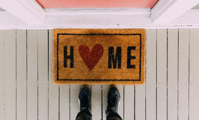 hand-held photo pointing downward, showing legs and feet standing on wooden deck in front of a "home" welcome mat with a heart, and the bottom edge of an orange door in a pink frame