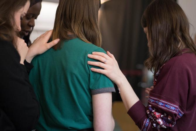 person in green shirt facing away from us, with three other people standing near them with their hands on the person's shoulders