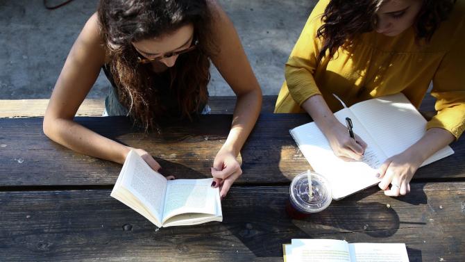 two people studying together at a wooden table, reading a book and taking notes