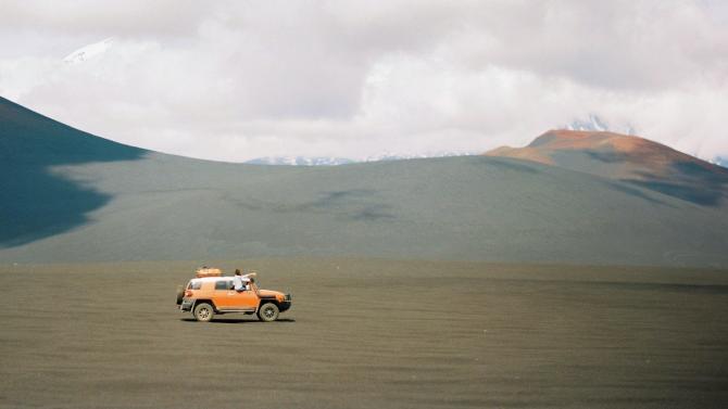 orange jeep driving through desert with luggage carrier on top and passenger sitting halfway out of a window, pointing ahead