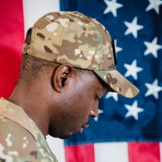 Black man in military uniform and cap standing in profile against American flag