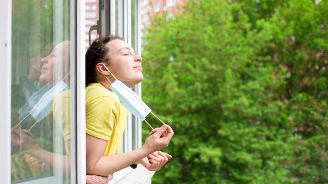 person leaning out a window into a tree-line space, removing paper surgical mask from their face