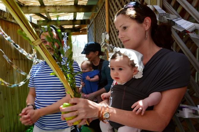 woman with baby in front carrier holding lulav and etrog in sukkah