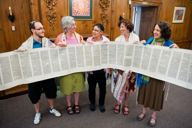 five people in tallitot holding a Torah scroll rolled open to display its text