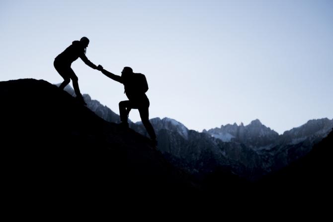 Silhouetted view of person climbing a rocky hill being helped up by someone already atop the hill