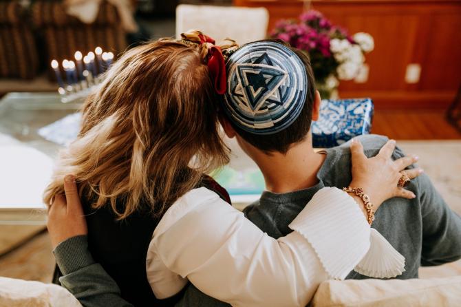 Long-haired and short-haired couple facing a table with a lit Hanukkah menorah