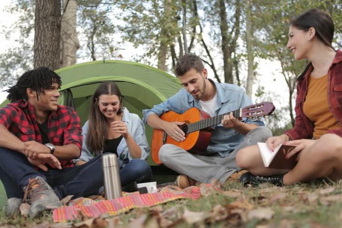 Group of adults seated on ground outside tent around a picnic blanket, singing with a guitar