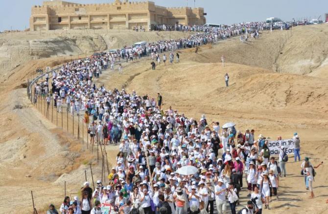 Massive march of women stretching into the distance through desert with ancient monastery in background