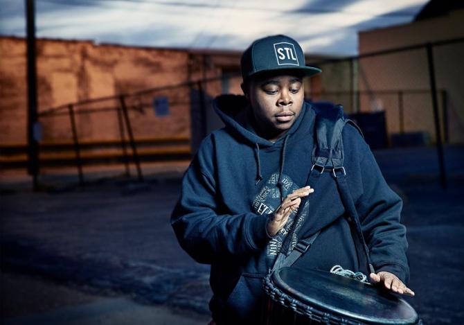 man in sweatshirt and St. Louis cap playing drum against desolate background