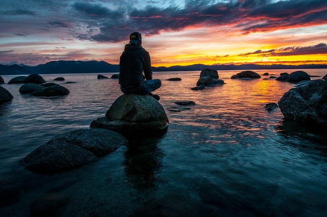 Silhouetted figure sitting on beach rocks watching sunset over lake