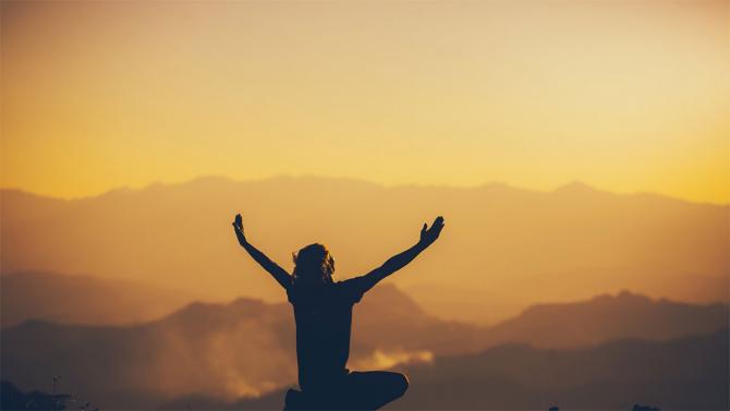 silhouetted person sitting in front of hills with hands raised