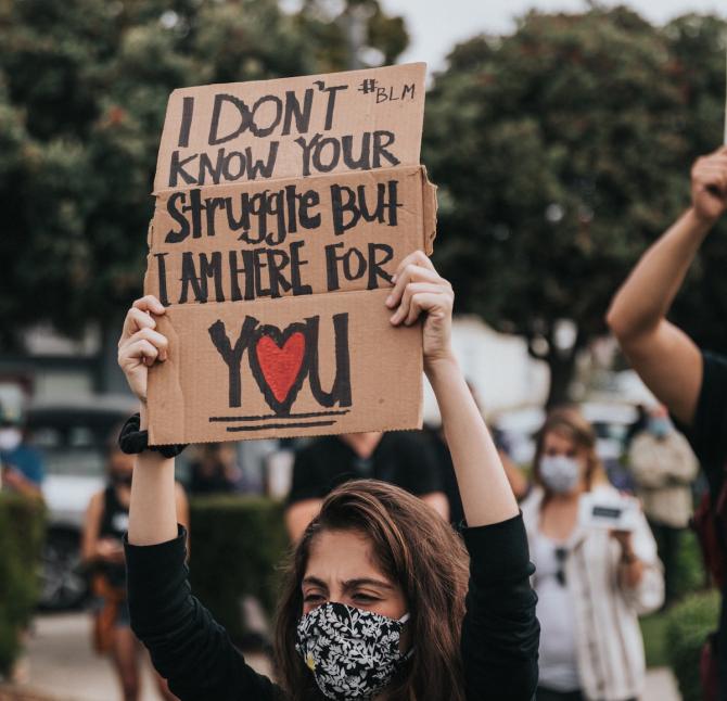 Masked woman at protest holding sign that reads "I don't know your struggle but I am here for you" and BLM