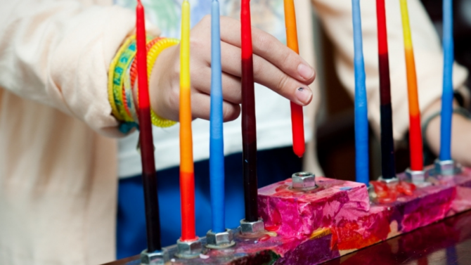 A hand holding a colorful candle above a colorful menorah