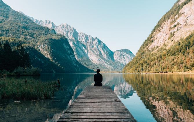 silhouette figure sitting at the end of a wooden deck facing a reflective lake with mountains on all sides