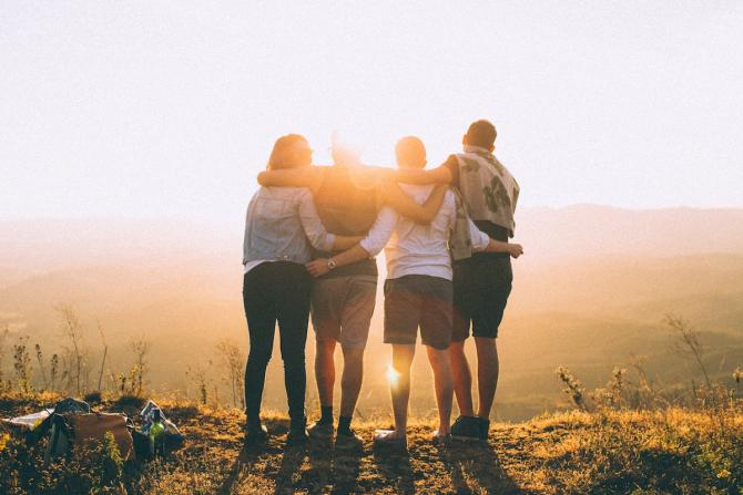 View from behind of four people on hilltop facing sunrise, arms around each other