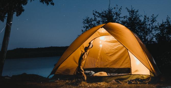 tent glowing brightly from lantern hung within, by a wooded lake under a starry night sky