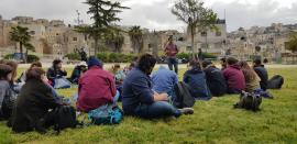 standing man speaking to a group of people seated on the grass in front of him. Buildings with Jerusalem stone and olive trees stand in the background