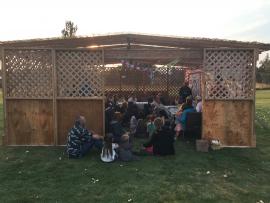 People gathered in a wooden sukkah with mountains faintly visible in the background
