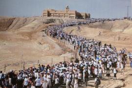 large march of women along desert road leading up to a building atop ahill