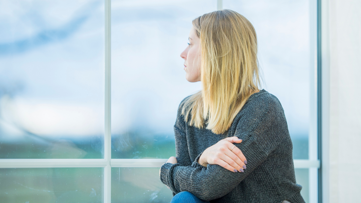 A blonde woman in a gray sweater looking somberly out a window