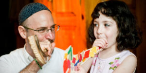A man and a young girl blowing colorful shofars indoors, smiling and looking at each other.