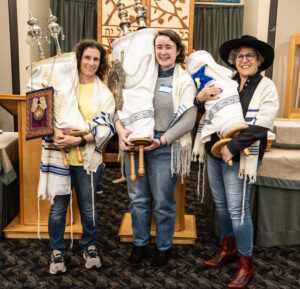 Three women smiling, each holding a Torah scroll, standing together in a synagogue setting.