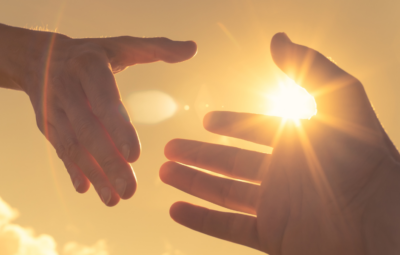 Two hands reaching toward each other against a bright sunlit sky.