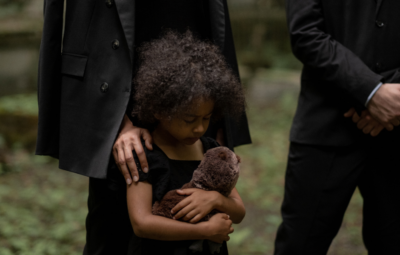 A child holding a stuffed animal stands between two adults in black outfits, likely at a somber outdoor event.