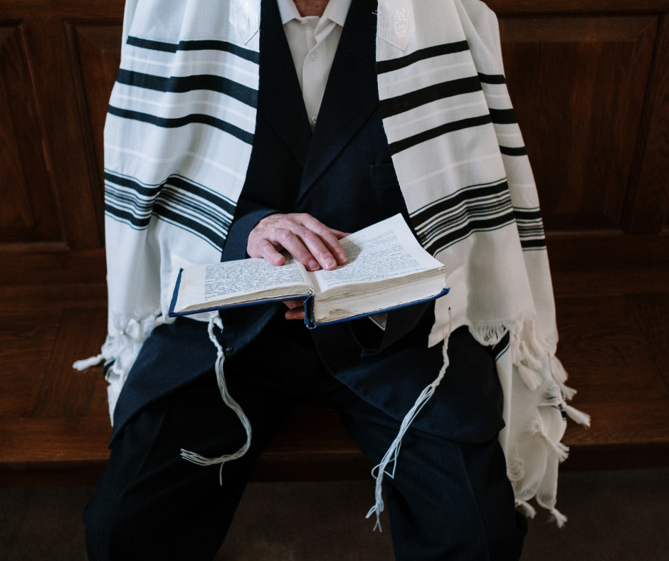Person wearing a prayer shawl, holding an open book and sitting on a wooden bench.