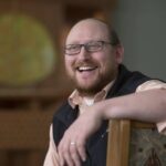 A bearded man with glasses and a smile sits casually in a chair, wearing a vest over a shirt.