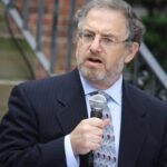A man in a suit and tie speaks into a microphone outdoors, with stairs and a railing in the background.