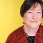 Smiling woman with short hair, wearing a red shawl and beaded necklace, against a yellow background.