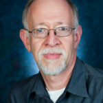 A man with glasses and a gray beard, wearing a dark shirt, against a mottled blue background.