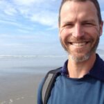 A man with a beard smiling on a beach, wearing a blue shirt and backpack. Ocean and cloudy sky in the background.