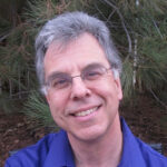 Smiling man with glasses and gray hair, wearing a blue shirt, standing in front of trees.