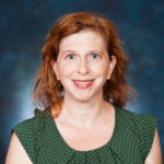 A person with red hair smiles at the camera against a blue background, wearing a green polka dot top.