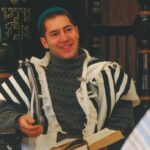 A person wearing a blue cap and striped shawl smiles while sitting indoors with an open book.