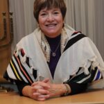 Person wearing a prayer shawl, smiling, standing indoors near a menorah.