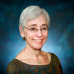 Smiling person with short gray hair and glasses, wearing a patterned top, against a gradient blue background.