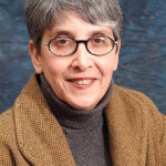 Person with short gray hair, glasses, and a brown jacket smiling against a textured blue background.