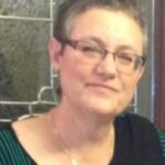 Person with short hair and glasses, wearing a black top with green stripes, standing in front of a paneled window.