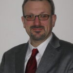 A man in a gray suit and red tie, wearing glasses, smiles against a plain background.