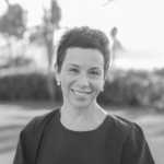 A person smiling outdoors with short hair, wearing earrings and a dark top, blurred trees in the background.