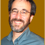 Smiling man with glasses, beard, and dark hair wearing a gray shirt against a brown background.