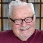 Elderly man with white hair, glasses, and a burgundy shirt smiling in front of a windowed background.