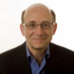 Man with glasses, bald head and wearing a suit, smiles at the camera against a plain background.